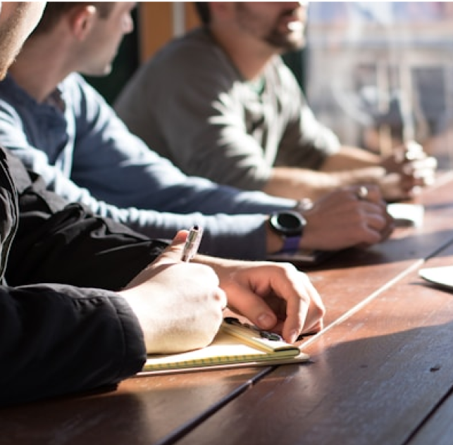 People sitting at a table, one person writing in a notebook